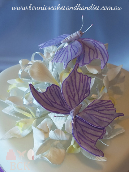 Purple butterfly wedding cake for a Mackay wedding:- traditional fruit cake with royal purple ribbon, white puffed hearts, butterflies, diamontes, white rosebuds, white Singapore orchids, two large purple butterflies - Bonnie's Cakes & Kandies, Gympie, Rainbow Beach & Sunshine Coast wedding cake decorator. The top tier was a heart shape, with the back of the heart raised approximately 2 inches, and the point of the heart almost touching the bottom tier, which was a square shape.